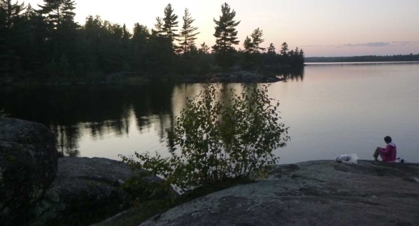 A person sits on the shore of a body of water. The sky appears in gentle colors as the sun either sets or rises. 